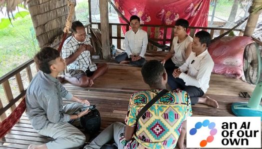 A group of men sits in a circle, in a wooden, open air pavilion, with an old electric fan on the right. There is a symbol with the words, an ai of our own.